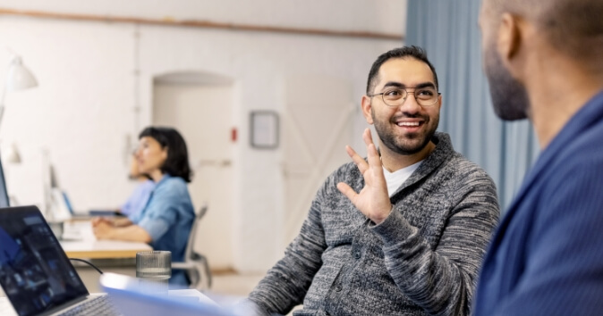 A smiling man wearing glasses is engaged in conversation, discussing what makes a good team, while sitting at a desk in an office setting. He is gesturing with his hand, indicating he is speaking to a colleague seated across from him. In the background, another person is working at a desk, and the environment appears bright and open with soft lighting.
