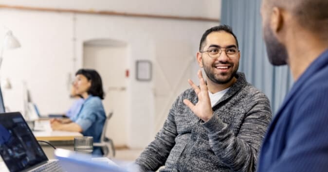 A smiling man wearing glasses is engaged in conversation, discussing what makes a good team, while sitting at a desk in an office setting. He is gesturing with his hand, indicating he is speaking to a colleague seated across from him. In the background, another person is working at a desk, and the environment appears bright and open with soft lighting.