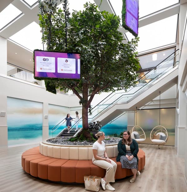 Workhuman's office interior featuring a large tree in the center, surrounded by a circular seating area with plush, multi-colored cushions. Two women are seated on the seating area engaged in conversation. Above the tree, two screens display information about individuals, suggesting a recognition or announcement theme. In the background, there are stairs leading to another level and additional people walking, creating a lively atmosphere. The walls are adorned with artistic murals that enhance the space's aesthetic.