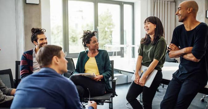 Two people standing, and others are sitting and talking and laughing in the workplace