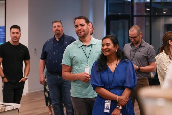 A group of six people stands in a brightly lit office space, smiling and chatting. One man in a light green shirt is sharing a laugh with a woman in a blue dress, while another man in a dark shirt observes in the background. All individuals appear engaged and friendly, suggesting a positive atmosphere. The interior features large windows and a modern design.