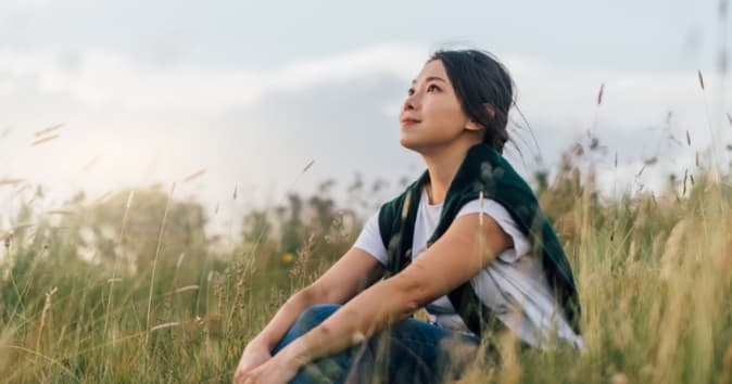 A woman sitting contemplatively in a field, exemplifying how rest is productive, as she gazes at the sky.