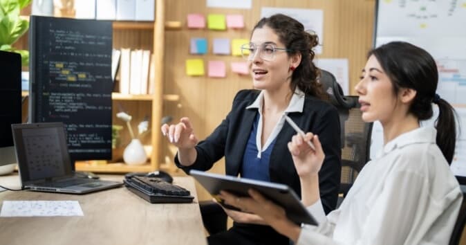 Two women are engaged in a discussion at a desk in a modern office environment. One woman, wearing glasses and a dark blazer, is animatedly speaking, using hand gestures to emphasize her points. The other woman, seated beside her in a white shirt, is holding a tablet and listening attentively. A laptop is open in front of them, and the background features a wall with organized notes and documents, conveying a collaborative workspace atmosphere.