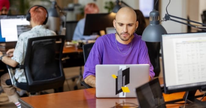 A focused person with a shaved head and earrings is working on a laptop at a modern office desk. He is wearing a purple shirt and appears to be engaged in his task. In the background, other individuals can be seen at their desks, contributing to a collaborative workspace atmosphere. The office has a bright and contemporary design with warm lighting.
