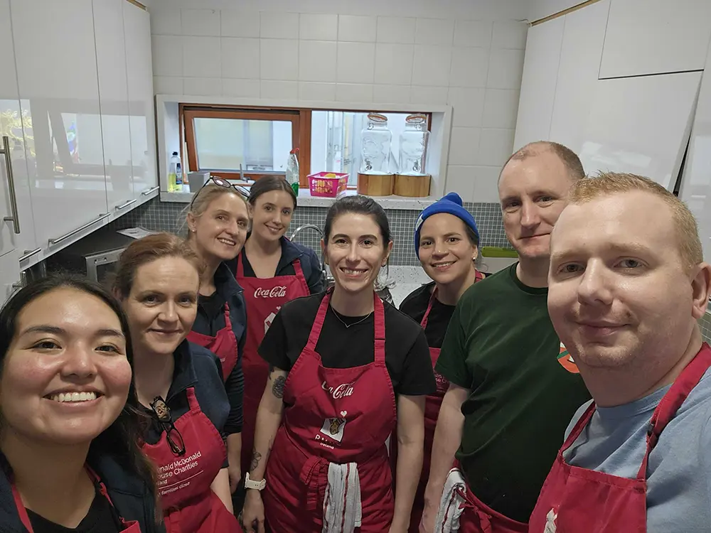A group of 8 volunteers from Workhuman are smiling and posing together in a kitchen setting, wearing matching red aprons.