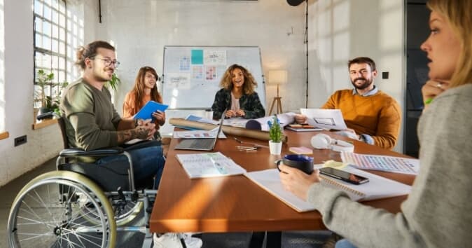 Group of people at a table celebrating Disability Pride Month, with the focus on a man in a wheelchair.