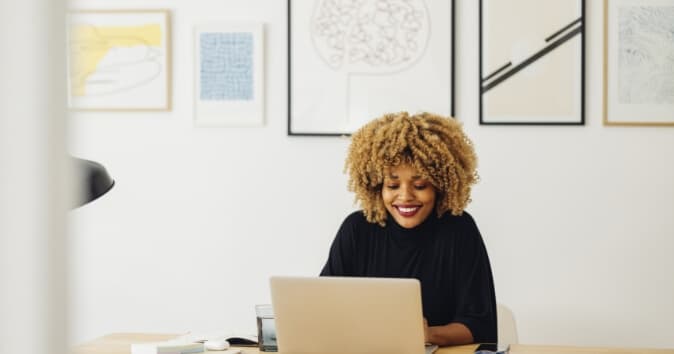 A woman with curly hair smiles while working on a laptop at a desk. The background features various framed artworks on the wall, and a glass of water is placed on the desk alongside a few desk accessories.