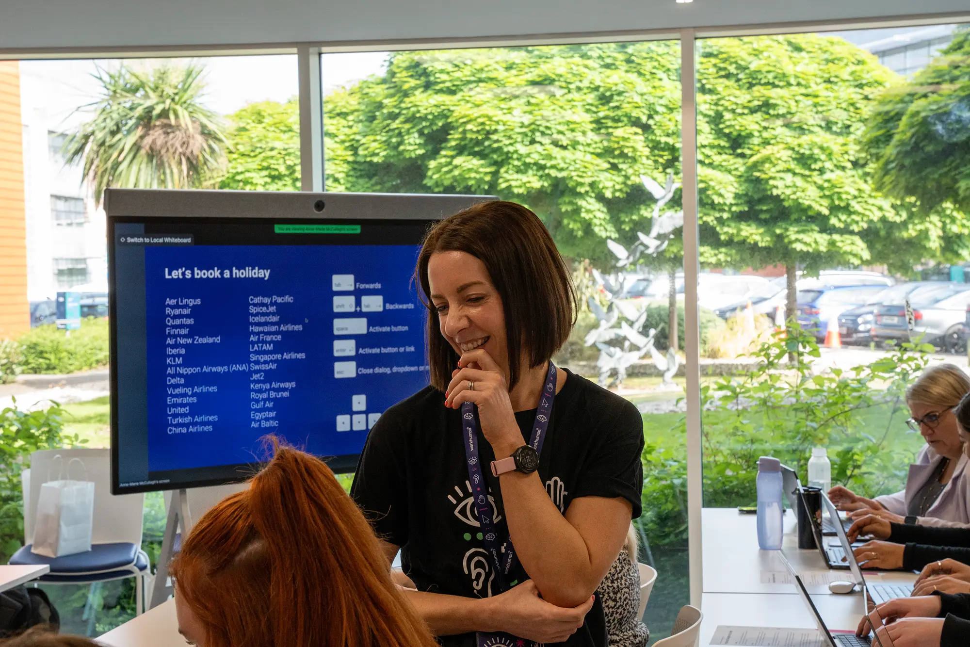 A woman with shoulder-length brown hair smiles while standing in front of a large screen displaying a booking interface titled "Let's book a holiday," which lists various airlines. In the background, two individuals are seated at a table, working on laptops, with greenery visible through a window behind them.