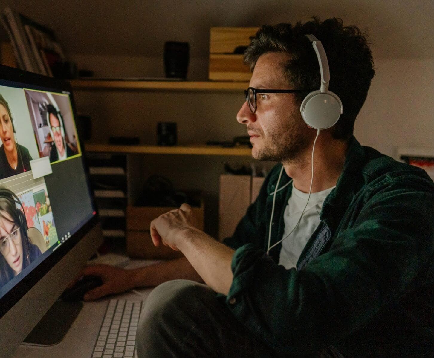 A person with curly hair and glasses is seated at a desk, wearing white headphones and looking at a large computer monitor displaying a video conference with multiple participants. The environment appears to be a dimly lit room with shelves and various items in the background.