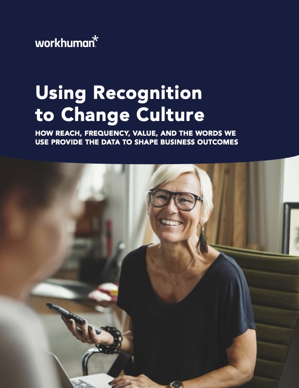 A smiling woman with glasses is sitting at a table, holding a smartphone. She is engaged in conversation with another person partially visible on the left. In the background, there are soft furnishings and a modern office setting. The image prominently features text that reads, "Using Recognition to Change Culture: HOW REACH, FREQUENCY, VALUE, AND THE WORDS WE USE PROVIDE THE DATA TO SHAPE BUSINESS OUTCOMES," along with the Workhuman logo.