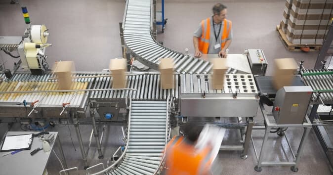 A busy warehouse setting with a conveyor belt system is seen in the image. Boxes are moving along the conveyor, while two workers in orange vests are engaged in the process. One worker is positioned near the conveyor, monitoring the boxes, while another is in motion nearby. The background features additional stacked boxes, indicating an active distribution environment.
