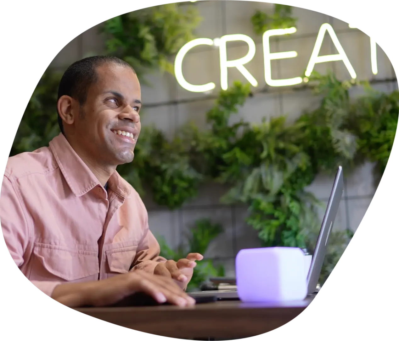 A smiling man wearing a salmon pink shirt sits at a laptop with his hands on the keyboard. Green planting spreads across the concrete wall behind him, and large illuminated letters spell create