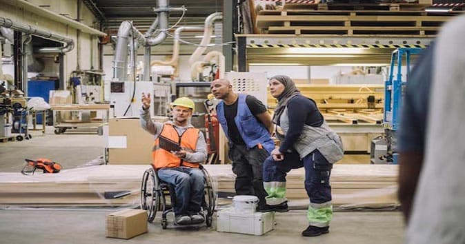 A disabled person sitting in a wheelchair wearing a vest, and safety helmet and instructing other employees