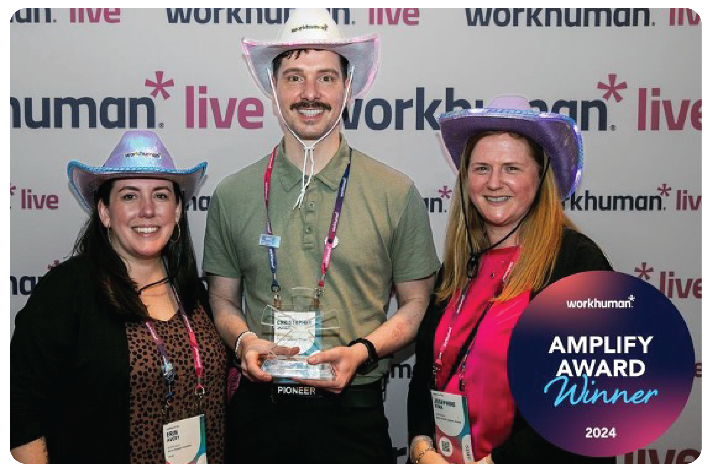 A group of three individuals stands in front of a workhuman.live backdrop, celebrating an Amplify Award for 2024. Each person is wearing a distinctive cowboy hat, with two in purple and one in blue. The central figure, holding an award, is flanked by two others: one on the left, smiling, and another on the right, also smiling. They exude happiness and excitement in this celebratory moment.