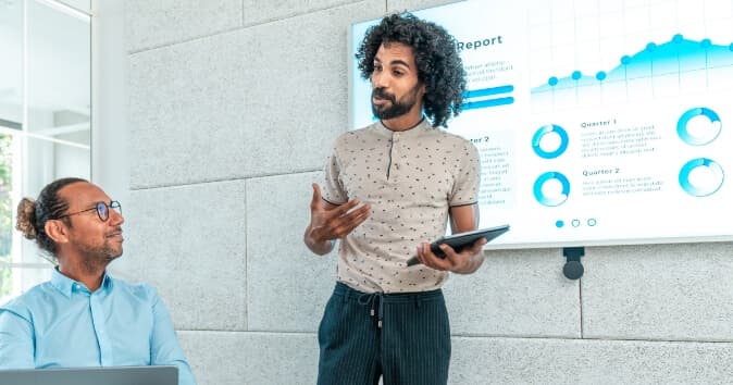A person with curly hair gestures while presenting a report in a modern office setting. They are holding a tablet and facing a seated colleague who appears engaged. A screen in the background displays a report titled "Quarter Report" with charts illustrating data trends for different quarters.