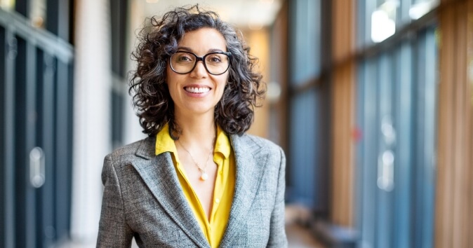 A smiling woman with curly hair is standing in a corridor, wearing a grey blazer over a yellow blouse. She has glasses and exudes a friendly demeanor, with wooden paneling and large glass windows visible in the background.
