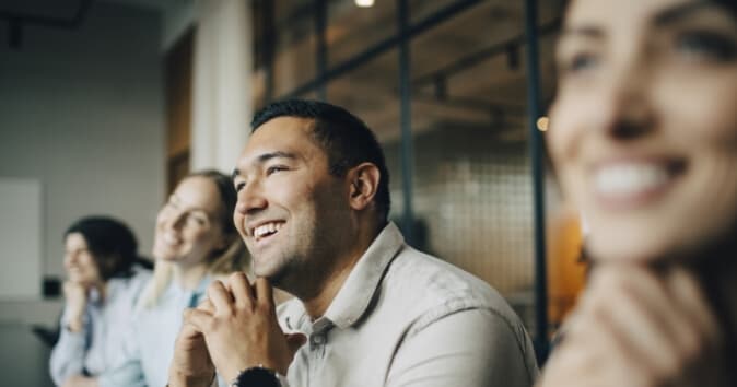 A group of four people sitting together, smiling and engaged in conversation. The focus is on a man in the foreground who is smiling and resting his hands under his chin. Two women are partially visible next to him, also smiling. In the background, another person is seated, contributing to a positive group dynamic in a modern office setting.