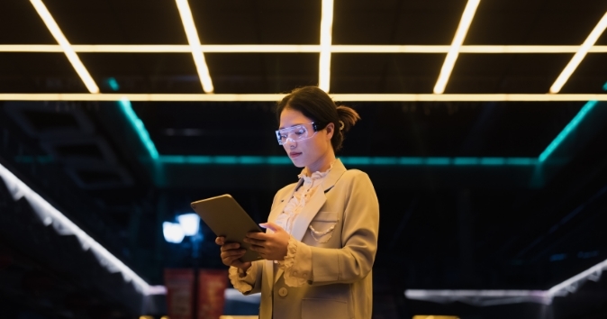 A woman wearing stylish eyewear and a light blazer is focused on a tablet she is holding. The setting features modern, bright lighting above her, creating a tech-savvy atmosphere.