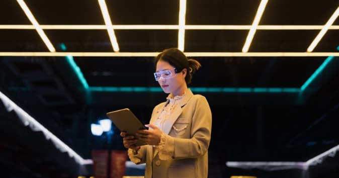 A woman wearing stylish eyewear and a light blazer is focused on a tablet she is holding. The setting features modern, bright lighting above her, creating a tech-savvy atmosphere.