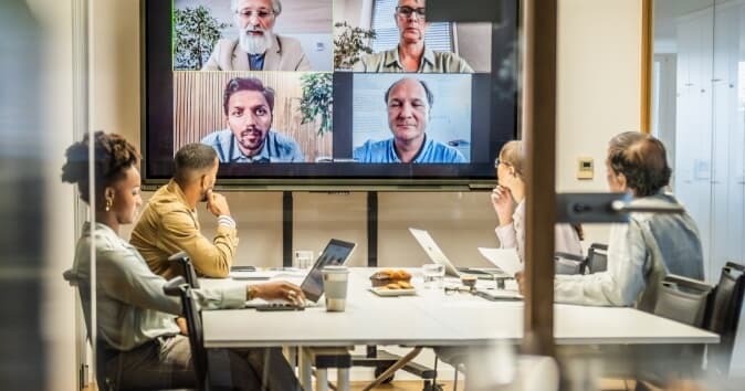 A group of professionals participates in a hybrid meeting. They are seated around a table with laptops, notepads, and a coffee cup visible. On a large screen in front of them, six individuals are shown in video conference, engaged in discussion. The setting has a modern office ambiance with neutral colors and ample natural light.