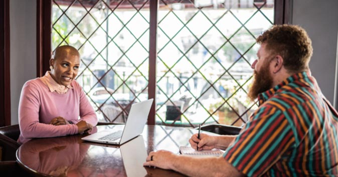 Women sitting in front of the laptop, and men taking notes, and they both are talking to each other