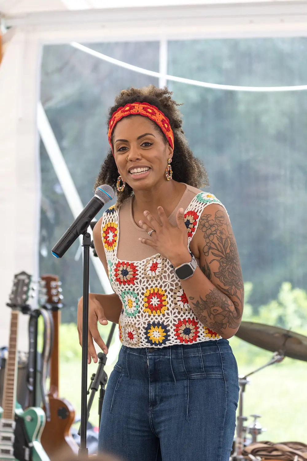 A woman with curly hair and wearing a colorful crocheted top stands at a microphone, smiling and engaging with an audience. She is gesturing with her hands and appears animated while speaking. In the background, there are musical instruments, including guitars, and a tent setting with natural light filtering through.