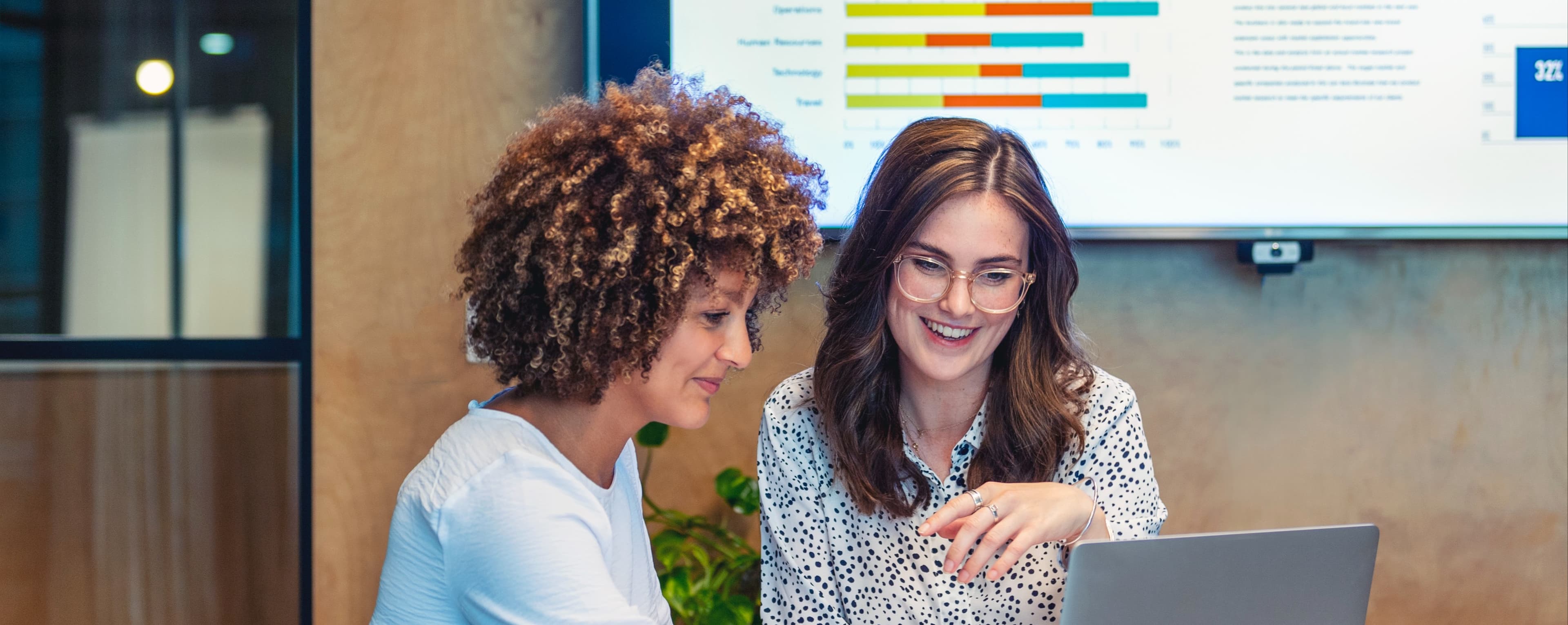 Two women are engaged in a discussion at a conference table, leaning towards a laptop in front of them. One woman, with curly hair, points to the screen while smiling, and the other, with straight hair and glasses, is also smiling and appears to be actively engaging in the conversation. Behind them, a monitor displays various graphs and data visualizations, indicating a professional setting. A coffee cup and some documents are visible on the table.