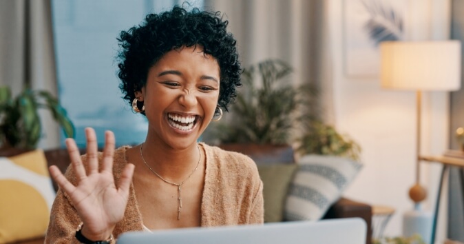 A woman waving and smiling towards a laptop, showcasing hybrid workplace culture.