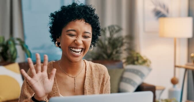A woman waving and smiling towards a laptop, showcasing hybrid workplace culture.