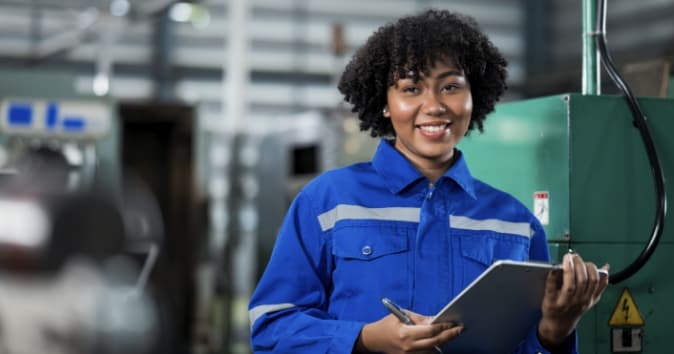 A smiling woman in a blue work uniform stands in a workshop, holding a clipboard in her right hand. The background features industrial machinery and equipment, highlighting a workplace environment.