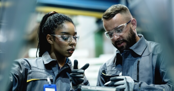 A female manager is showing a male employee how to operate an electronic device, demonstrating effective leadership. Both are wearing safety glasses and gloves. The woman has long, dark hair tied back and appears focused on the task, while the man, with a beard, points with one finger and looks at the device with interest. They are in an industrial setting with blurred machinery and tools in the background, suggesting a workshop or factory environment.