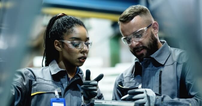 A female manager is showing a male employee how to operate an electronic device, demonstrating effective leadership. Both are wearing safety glasses and gloves. The woman has long, dark hair tied back and appears focused on the task, while the man, with a beard, points with one finger and looks at the device with interest. They are in an industrial setting with blurred machinery and tools in the background, suggesting a workshop or factory environment.