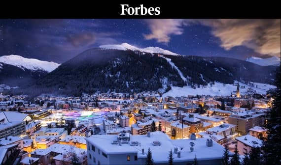 A panoramic view of a snowy town at night, with numerous buildings illuminated by lights beneath a clear sky filled with clouds. In the foreground, rooftops are visible, covered in snow, while in the background, snow-capped mountains rise against the night sky. The word "Forbes" is prominently displayed at the top of the image.