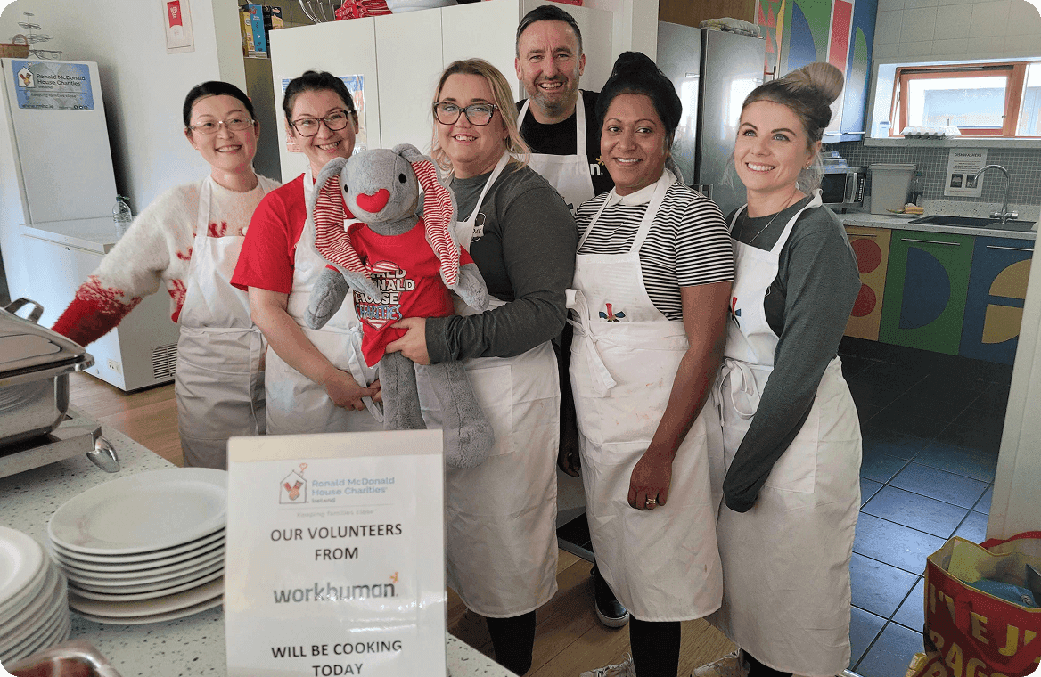 A group of six volunteers from Workhuman is smiling and posing together in a kitchen setting, wearing matching white aprons. One volunteer is holding a plush toy dressed in a red shirt with the Ronald McDonald House Charities logo. In the foreground, a sign reads, "OUR VOLUNTEERS FROM Workhuman WILL BE COOKING TODAY." The kitchen has a colorful backsplash, and various cooking supplies can be seen in the background.