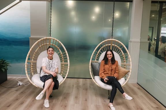 Two women are sitting in hanging chairs in a modern office space. The woman on the left is wearing a white top and black skirt, smiling at the camera, while the woman on the right is dressed in a brown blouse and black pants, also smiling. The background features frosted glass and a scenic wall mural.