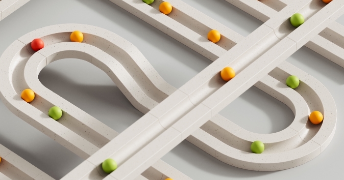 A top-down view of a maze-like track made from light-colored, curved and straight segments. Colorful balls in red, orange, yellow, green, and light green navigate the pathways, with some balls resting within the curves of the track while others are in motion. The background is a muted gray, enhancing the visibility of the vibrant balls and the track's design.