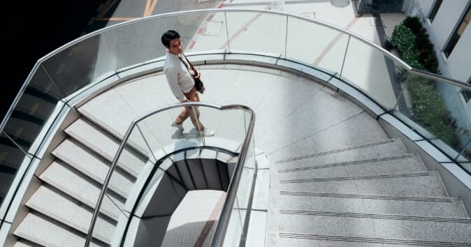 An employee walking down a spiral staircase in a well-lit space, with glass railings and a modern design. The employee, dressed in light attire, is seen from above as they approach the lower level of the staircase. It is meant to symbolize an employee on the path of their individual development plan.