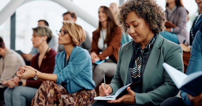 A diverse group of attendees sitting in a seminar or workshop setting, with one woman in the foreground taking notes in a notebook. She has curly hair and is wearing a teal blazer. Other attendees, both men and women, are engaged in the session, some looking ahead and others appearing thoughtful. The environment appears bright and modern.