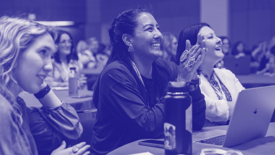 A group of diverse attendees at a conference are engaged in conversation, reflecting a lively atmosphere. In the foreground, a woman wearing a striped shirt is smiling and animatedly speaking with a companion, while several others around them are attentive and smiling, indicating camaraderie and excitement. The background is filled with people, some holding drinks, all contributing to the energetic environment of the event.