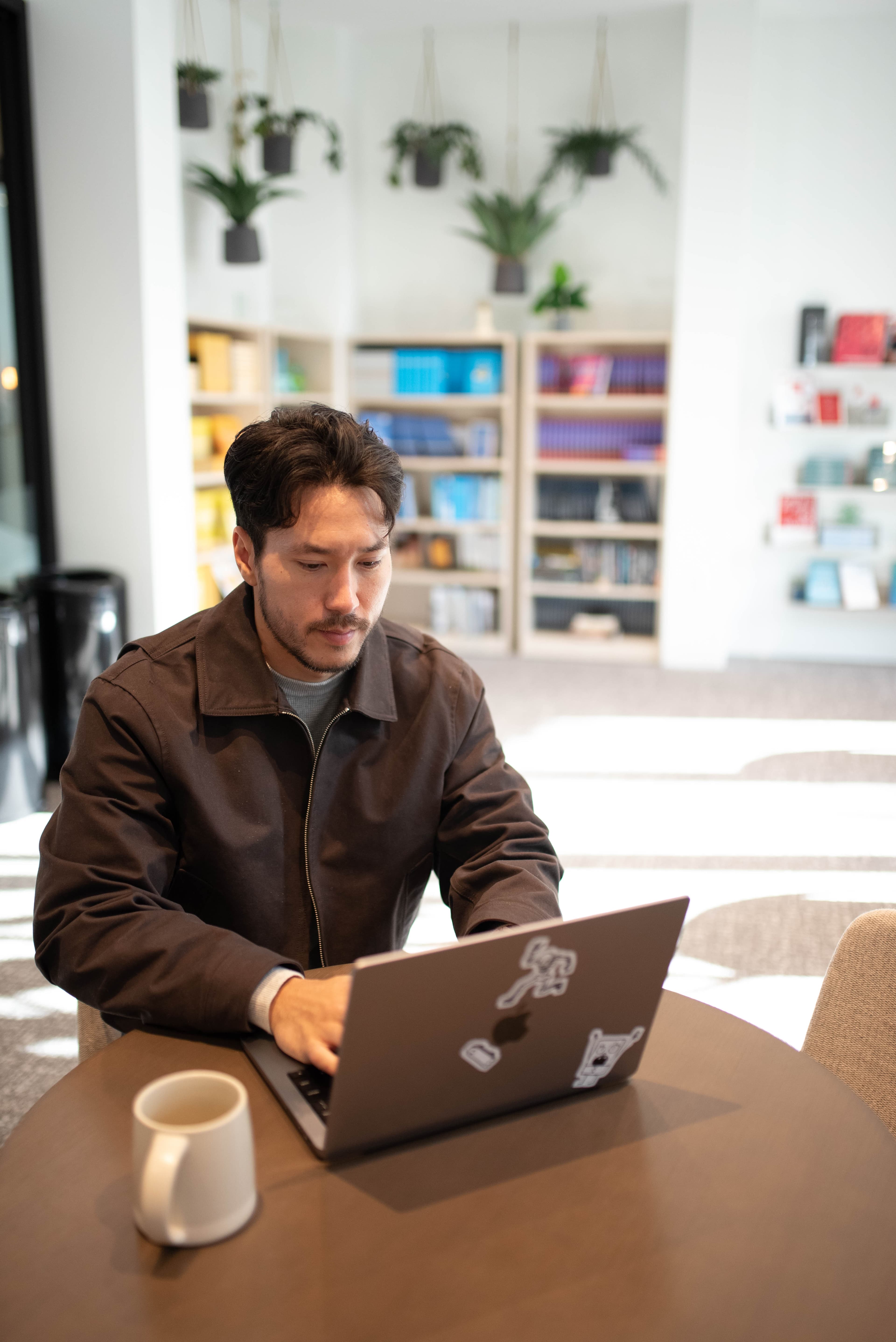 Man working on a laptop with stickers, seated at a round table in a modern library. Bookshelves and hanging plants create a focused, serene vibe.
