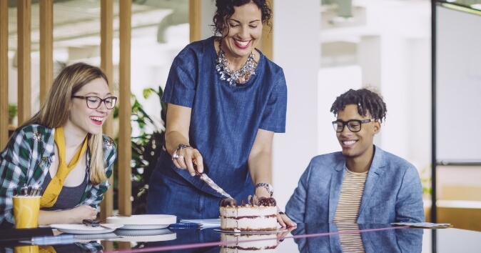 Three people in a room for a birthday, with a woman cutting a cake.