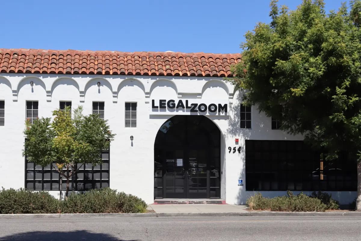 A white building with a red tile roof features large black letters spelling "LEGALZOOM" above the entrance. The façade includes several rectangular windows and an arched doorway, with the address "954" displayed next to the door. There are small shrubs and trees lining the front of the building, and the sky is clear and blue.