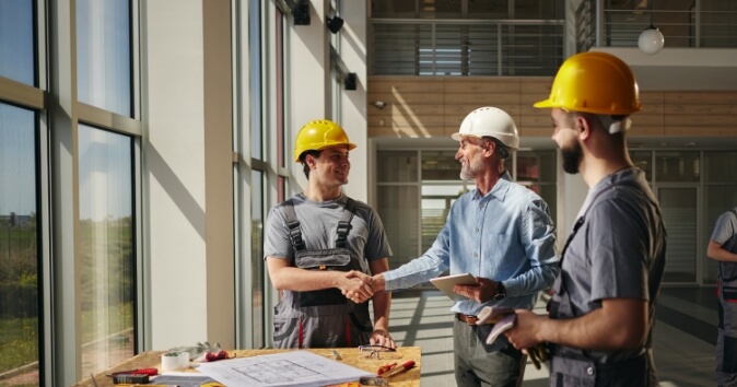 A construction worker in a yellow hard hat shakes hands with a man in a light blue shirt, while another worker in a gray hard hat observes. They are in a well-lit area with large windows displaying a green landscape outside. A table in front of them holds blueprints and tools, indicating a collaborative work environment. This scene subtly highlights the potential for favoritism in the workplace, where personal connections or preferences could influence professional interactions.