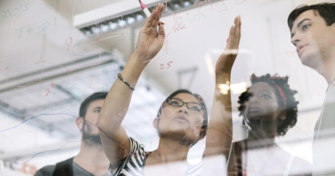 A diverse group of four people in a collaborative environment are engaged in a discussion, with one woman at the forefront using a marker to write on a transparent surface or whiteboard. The others are observing her, seemingly contributing to the conversation. The background includes various mathematical notations or diagrams that are faintly visible on the glass surface, indicating a brainstorming or planning session.