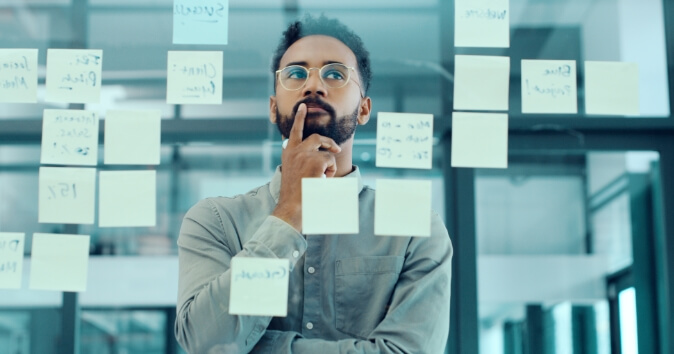 A man wearing glasses stands in front of a glass wall covered with sticky notes, deep in thought. He has a hand on his chin, contemplating the information displayed on the notes, which appear to contain various ideas or tasks. The environment suggests a collaborative workspace or meeting area, emphasizing a focus on planning or brainstorming.