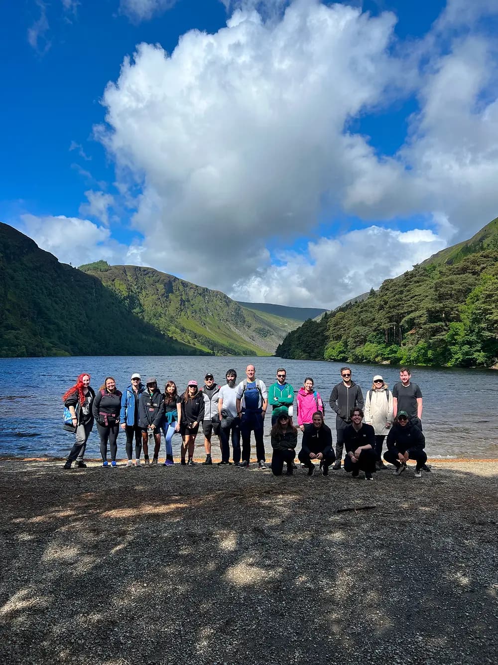 A group of diverse individuals stands together along the shore of a lake, with lush green mountains and a bright blue sky in the background. The scene depicts a vibrant outdoor setting, suggesting a day of teamwork or camaraderie in nature.
