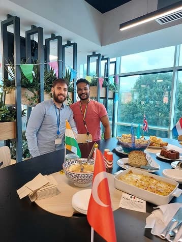 Two men are standing beside a table laden with various dishes and foods, decorated with plates displaying flags from different countries. The setting features large windows in the background, allowing natural light to illuminate the scene. The men are both smiling, wearing casual clothing, and appear to be enjoying the moment together amidst an inviting atmosphere filled with greenery.