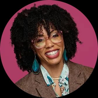 A woman with curly black hair and glasses smiles, wearing a colorful blouse and a brown blazer, against a pink background. Her expression is warm and approachable, exuding positivity.