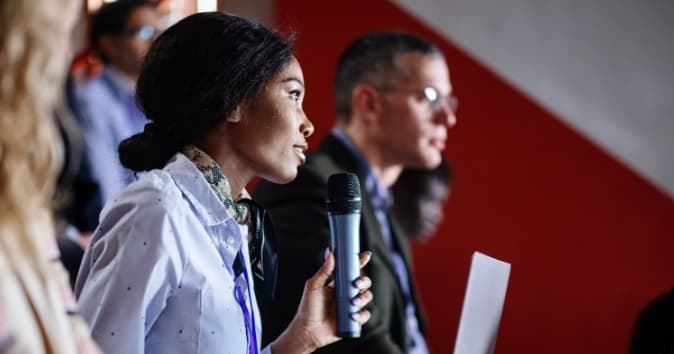 A woman is engaged in a discussion while holding a microphone, poised to ask a question or make a comment. She has long hair and is wearing a white shirt. Seated next to her is a man in glasses who appears attentive. The background is blurred, with hints of other attendees visible. The setting suggests a conference or panel discussion.