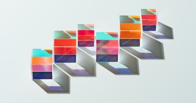 A series of colorful, transparent blocks arranged in a staggered formation, casting distinct shadows on a light background. The blocks feature various patterns and hues, including shades of red, blue, orange, and green, creating a vibrant and visually dynamic display.